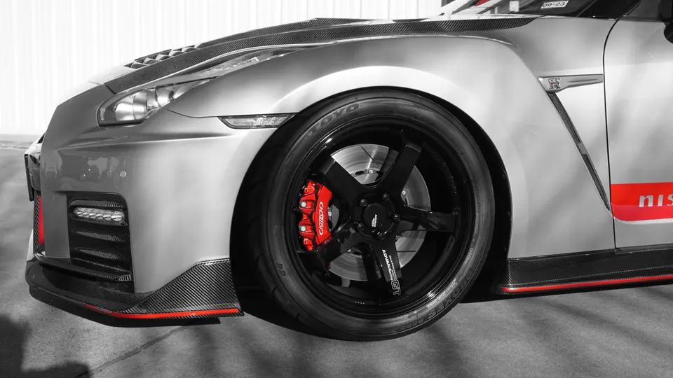 A close-up of a silver Nissan GT-R's front wheel, featuring black multi-spoke wheels, red brake calipers, and carbon fiber aero.