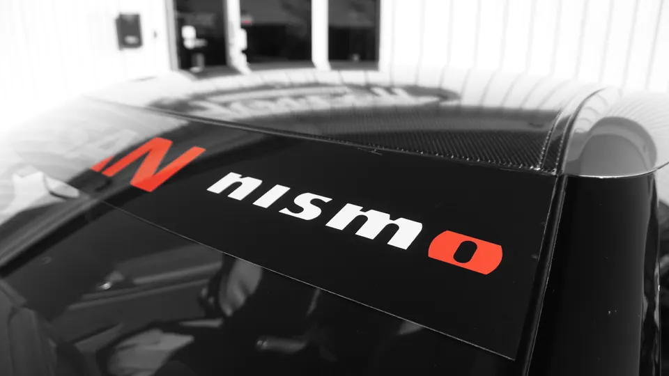 Close-up of a car's rear window featuring a black Nismo banner with white and red lettering, and a carbon fiber roof.