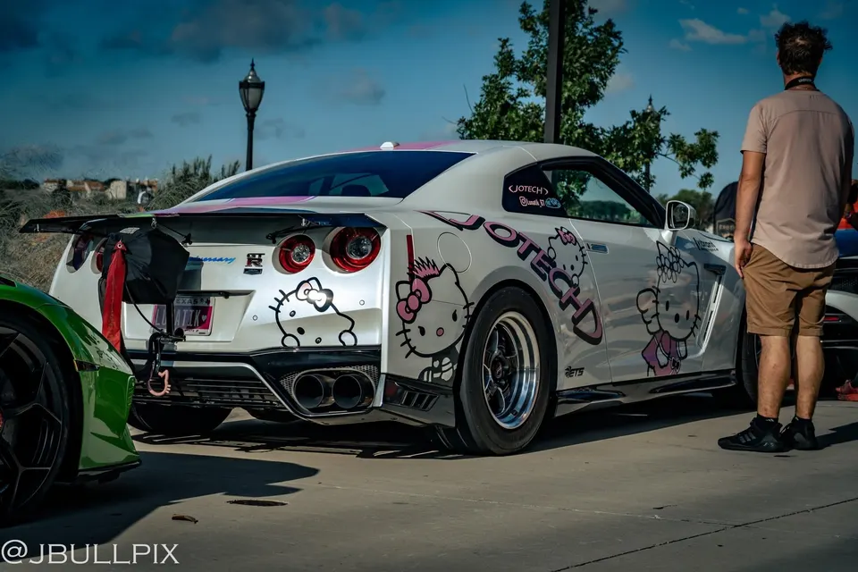 White Nissan GT-R with Hello Kitty decals and drag wheels at a car meet, featuring a large rear wing.