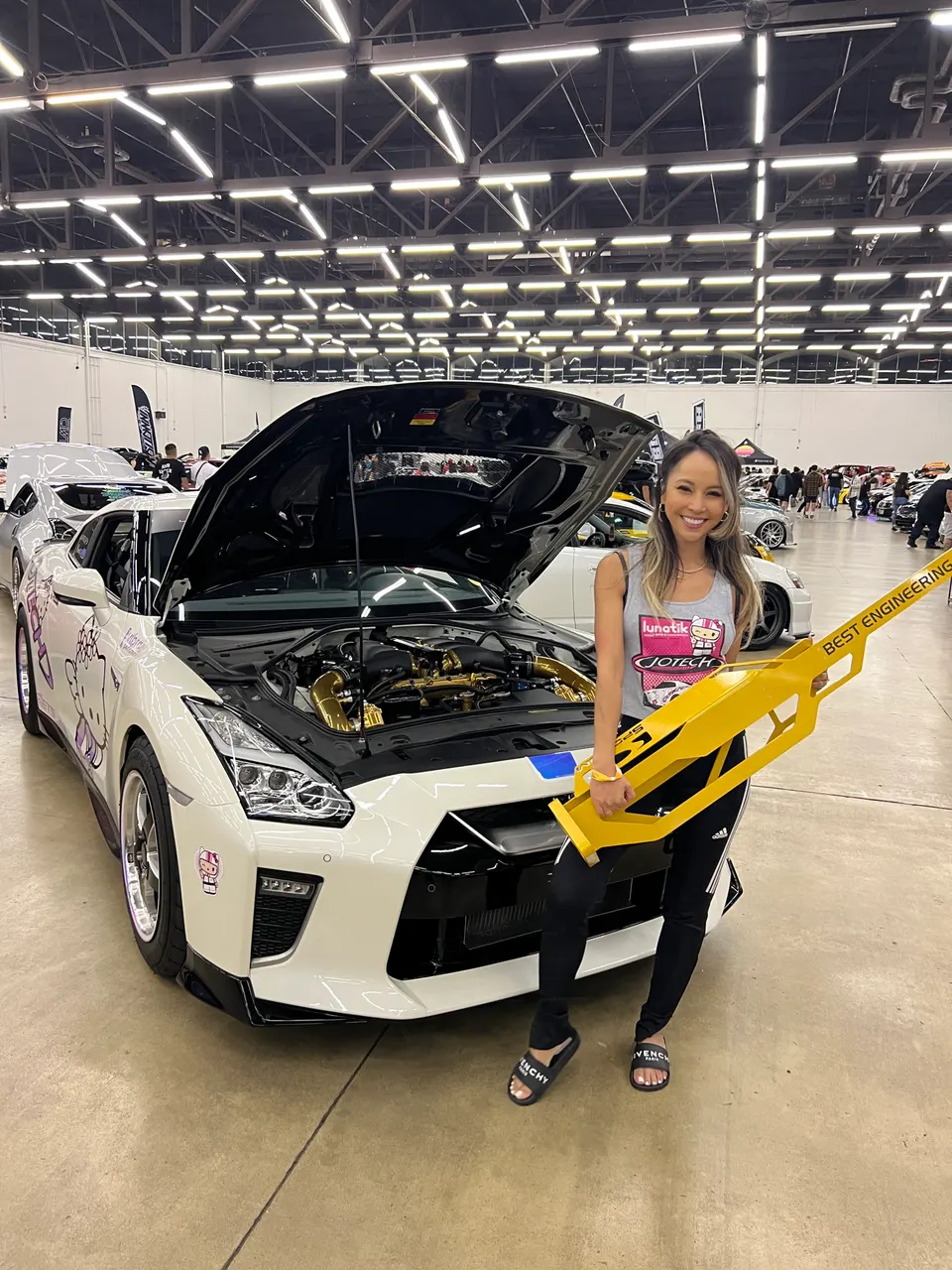 A woman stands next to a white Nissan GT-R with its hood open, showcasing a gold engine bay, at a car show.