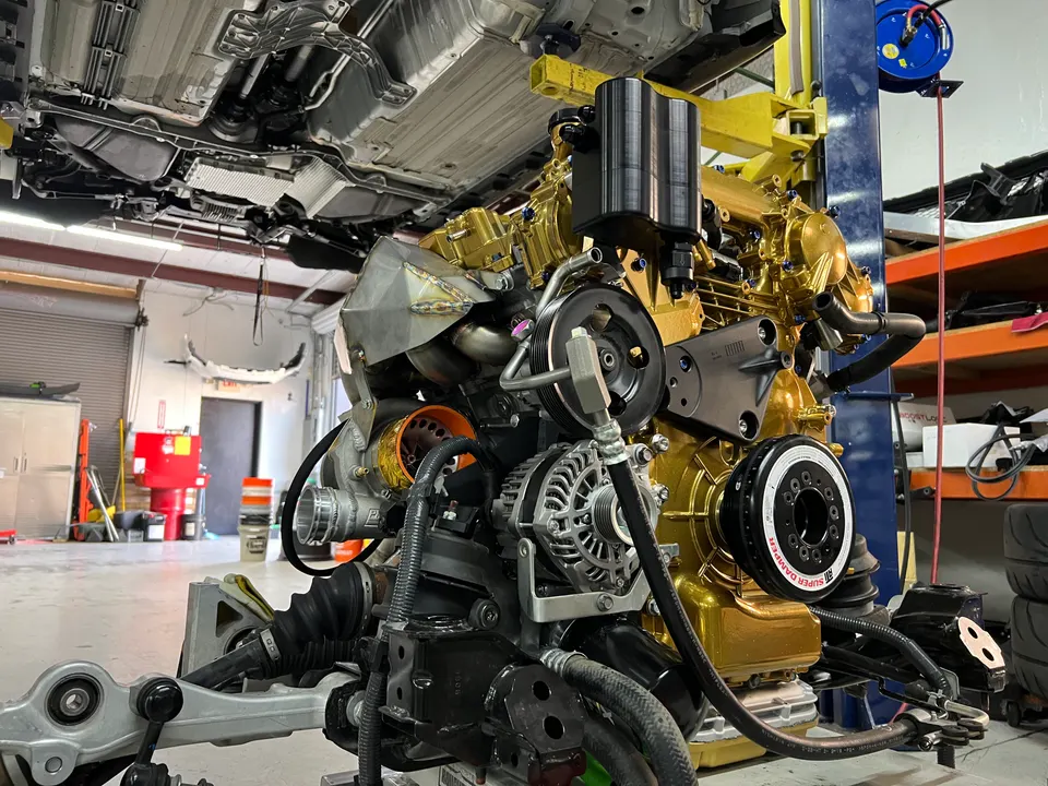 Highly modified gold-painted engine assembly with a large turbocharger in a performance tuning shop, with a car on a lift overhead.