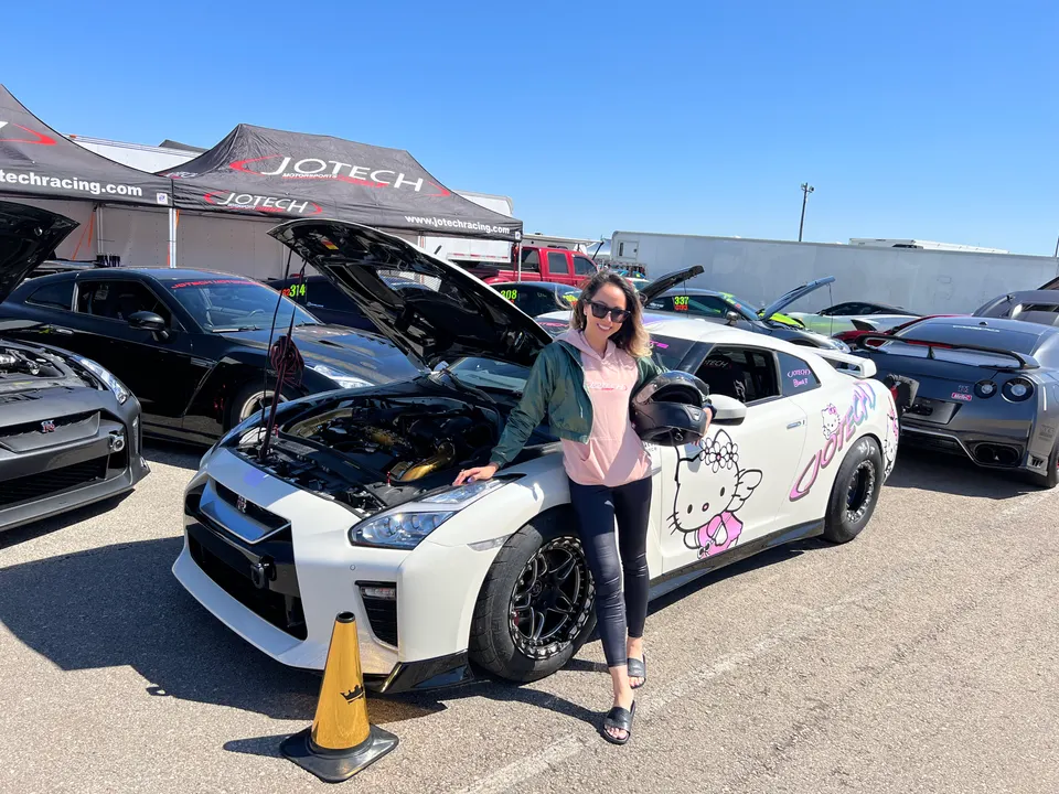 A woman in sunglasses and a jacket stands next to a white Nissan GT-R drag car with an open hood and Hello Kitty livery at a Jotech Motorsports track event.