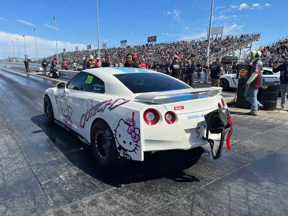White Nissan GT-R with Hello Kitty livery in the staging lane at a drag strip, with spectators in the background.