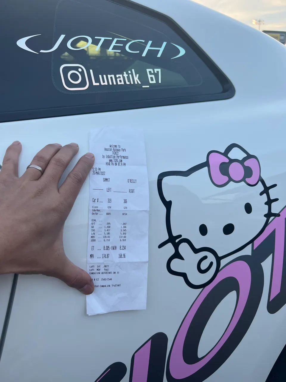 A hand holding a drag race timeslip against a white Nissan GT-R with Jotech and Hello Kitty decals.
