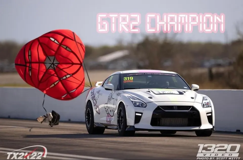 A white Nissan GT-R drag car with a deployed red parachute on a drag strip at TX2K22, with 'GTR2 CHAMPION' text.