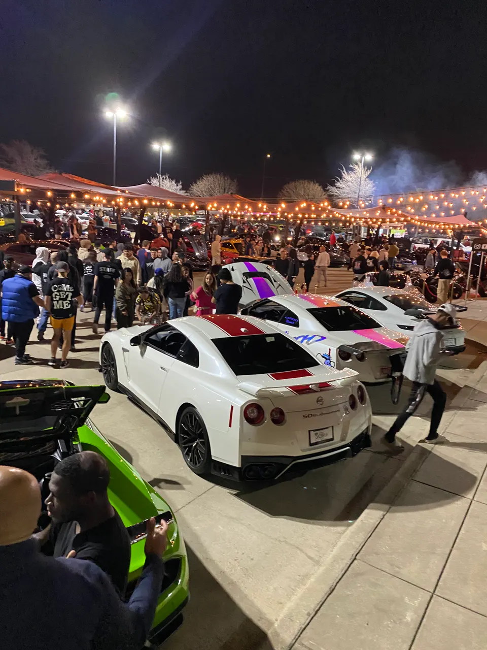 A night shot of a car meet with multiple modified cars and a crowd of people. A white Nissan GT-R is in the foreground.