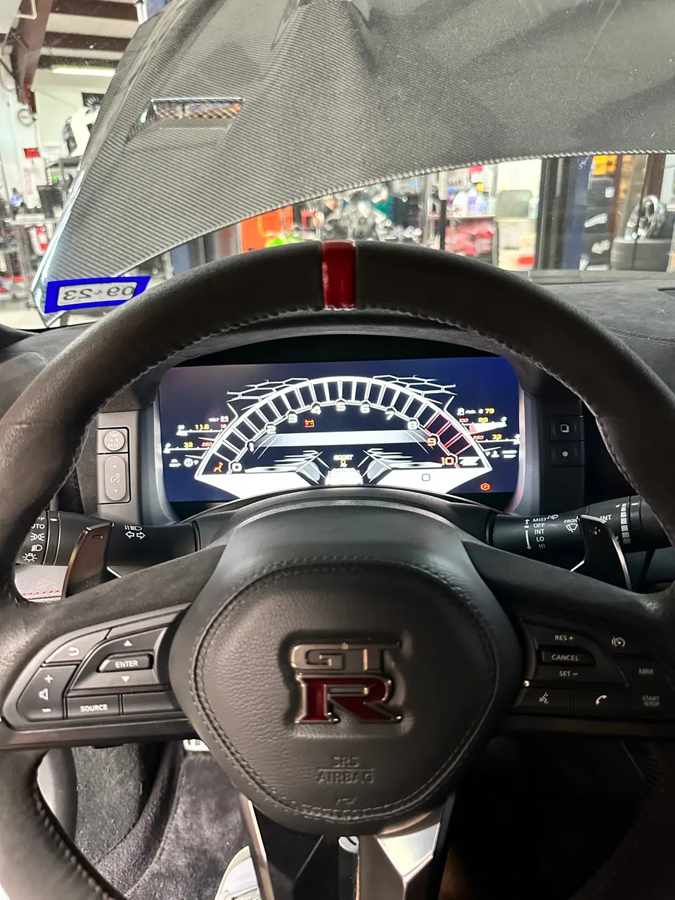 Nissan GT-R interior with custom steering wheel and digital dashboard displaying performance data, seen from a tuning shop.