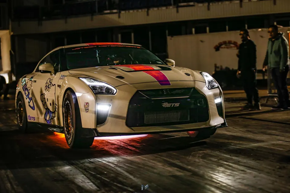 White Nissan GT-R drag car with Hello Kitty livery and underglow lighting at a night track event.