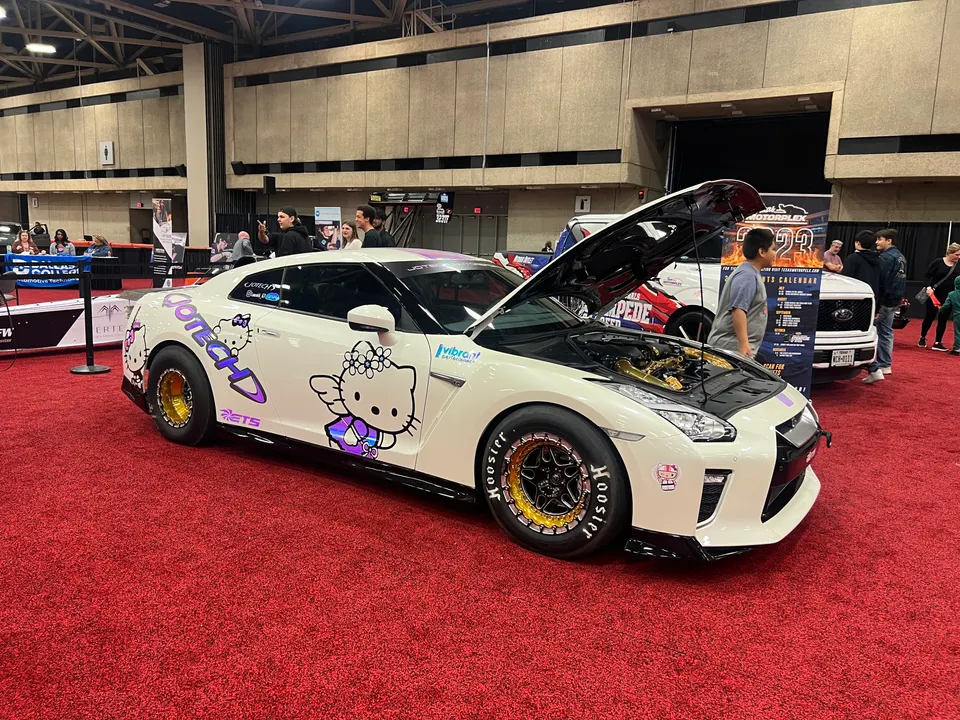 A white Nissan GT-R with Hello Kitty livery and drag wheels displayed at an indoor car show with its hood open.