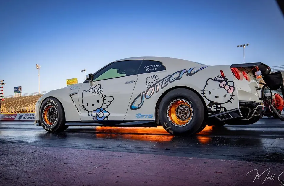 A white Nissan GT-R with Hello Kitty livery and Jotech branding performs a burnout on a drag strip with flames.