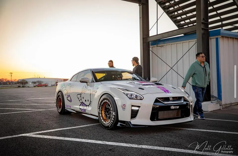 White Nissan GT-R with Hello Kitty livery and drag tires parked at a track facility during sunset.