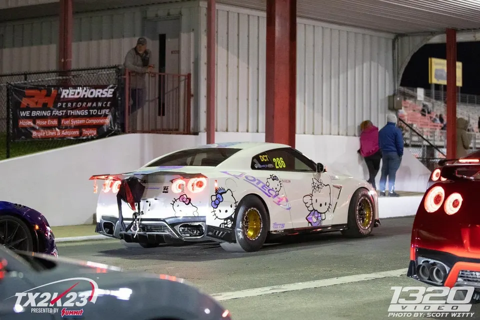 White Nissan GT-R with Hello Kitty decals and a drag parachute at a nighttime drag strip event.