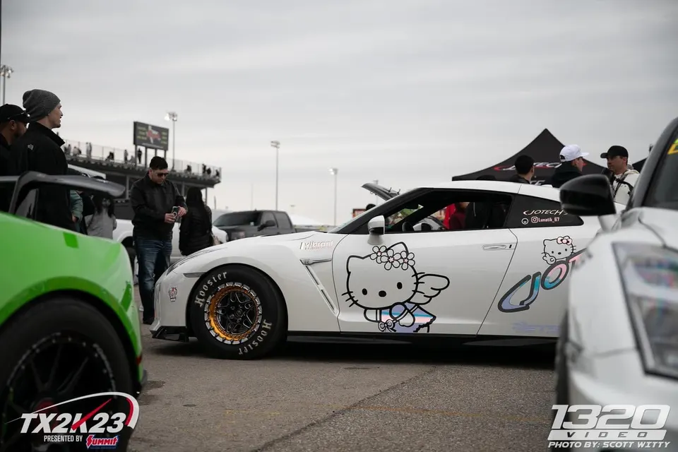 White Nissan GT-R with Hello Kitty decals and drag slicks at a track event with spectators.
