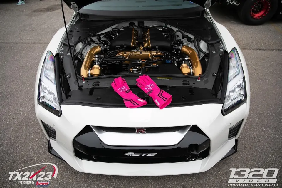 A white Nissan GT-R with its hood open, showcasing a custom gold engine bay with boost pipes and blow-off valves. Pink racing gloves rest on the front bumper.
