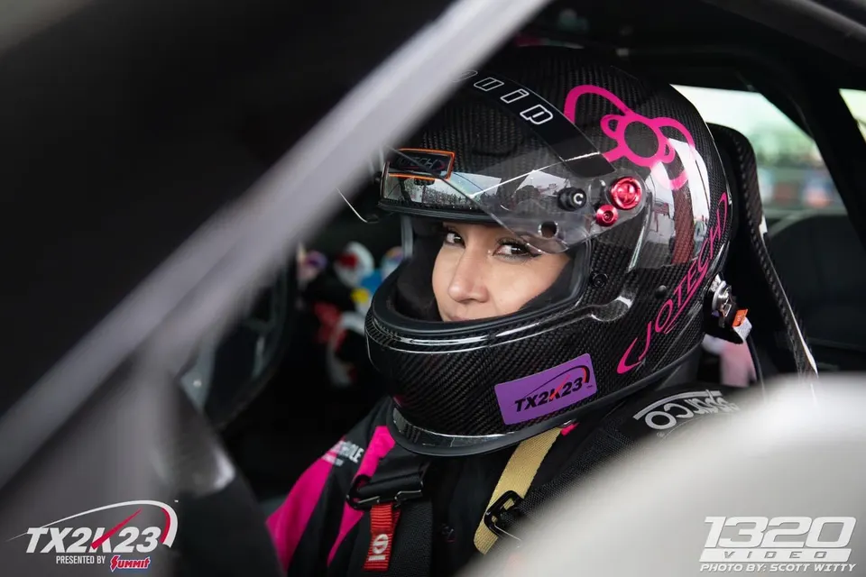 Female race car driver in a carbon fiber helmet with pink accents and a racing suit inside a car at TX2K23.