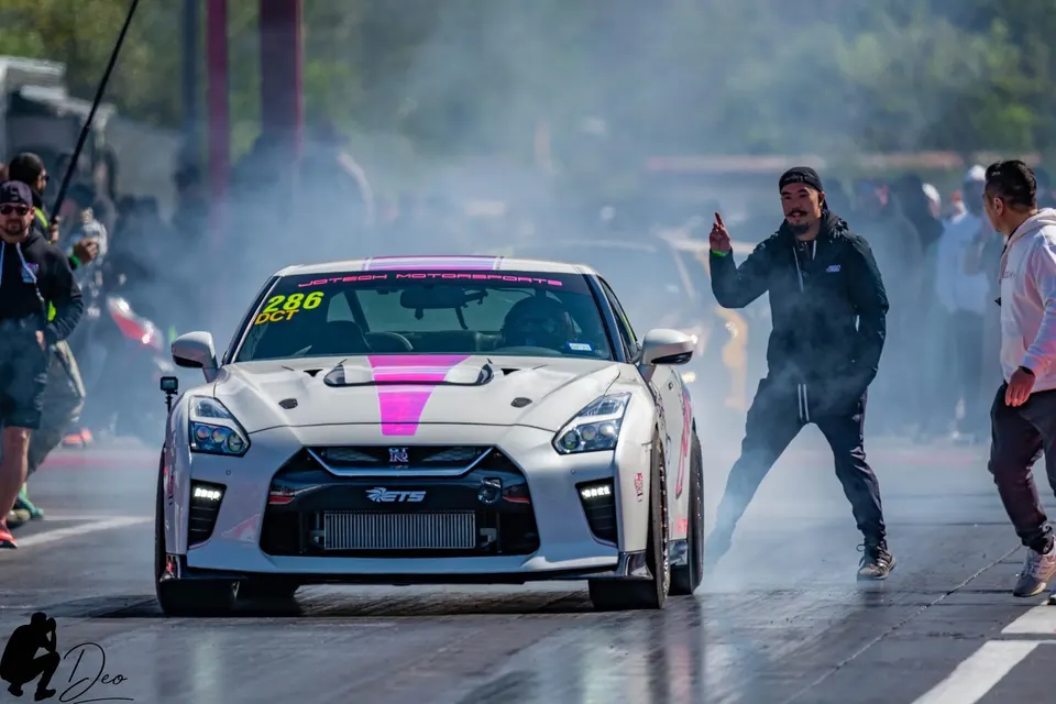 A white Nissan GT-R with pink accents performs a burnout at a drag strip, with a crew member signaling.