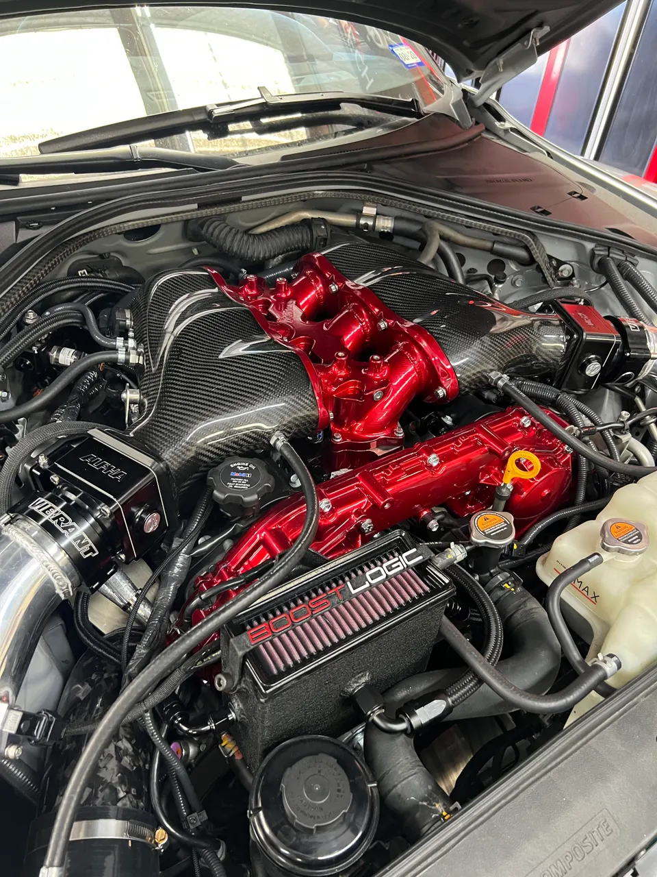 Close-up of a high-performance engine bay featuring a carbon fiber intake manifold, red valve covers, and aftermarket induction components from Boost Logic, Vib