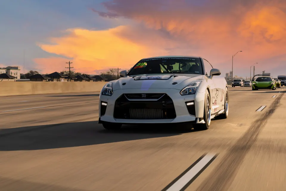 A white Nissan GT-R with an aftermarket intercooler drives on a highway at sunset, with other cars in the background.