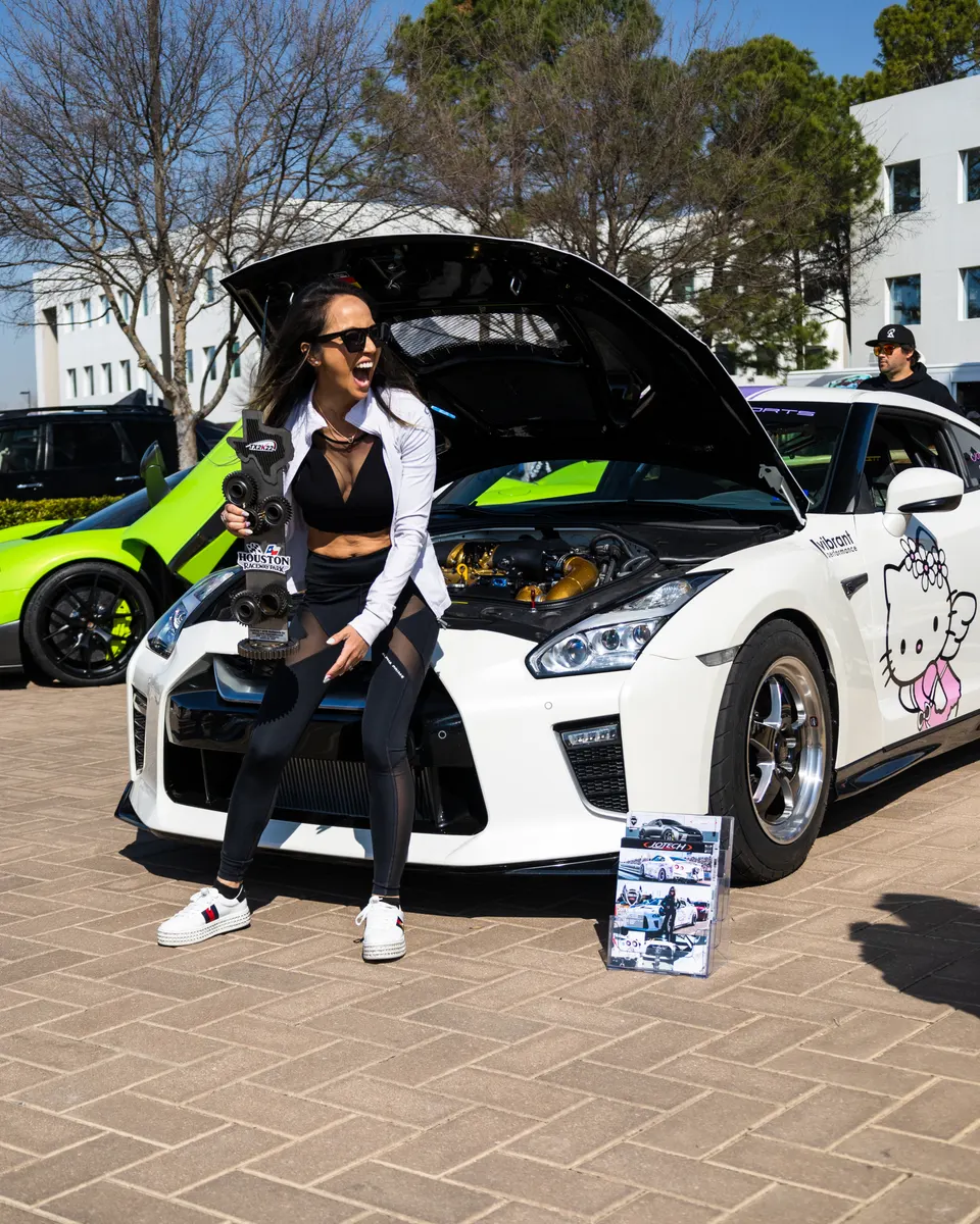 A woman in sunglasses and activewear poses with a white Nissan GT-R with an open hood and Hello Kitty decal at an outdoor car show.