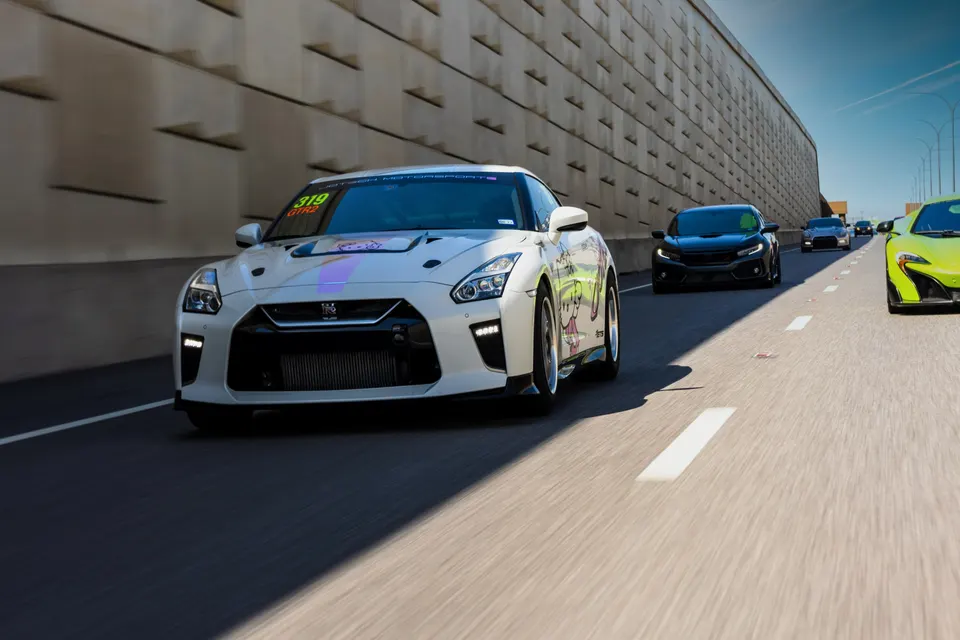 A white Nissan GT-R with custom livery leads other performance cars like a black Honda Civic Type R and a green McLaren on a highway.