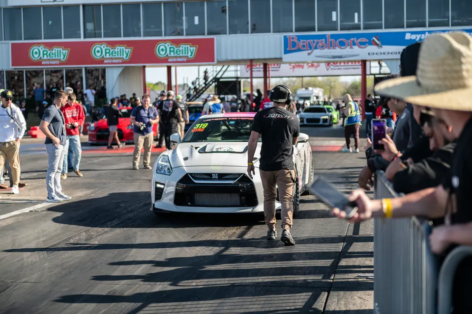 A white Nissan GT-R race car in the staging lanes of a drag strip, with a Jotech Motorsports crew member walking in front.