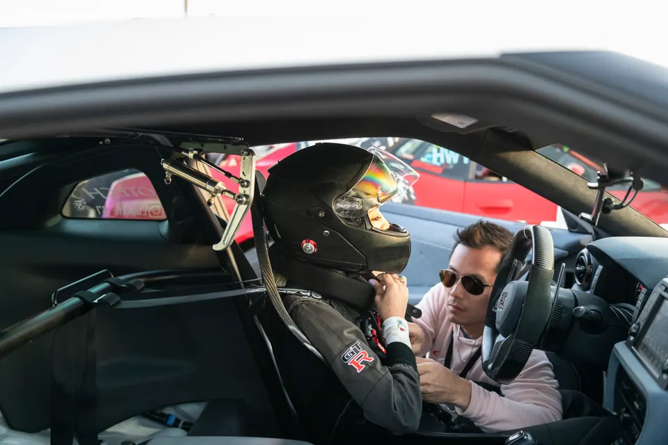 A person in a racing helmet and suit, with a GT-R logo, sits in the driver's seat of a car with a roll cage, while another person assists.