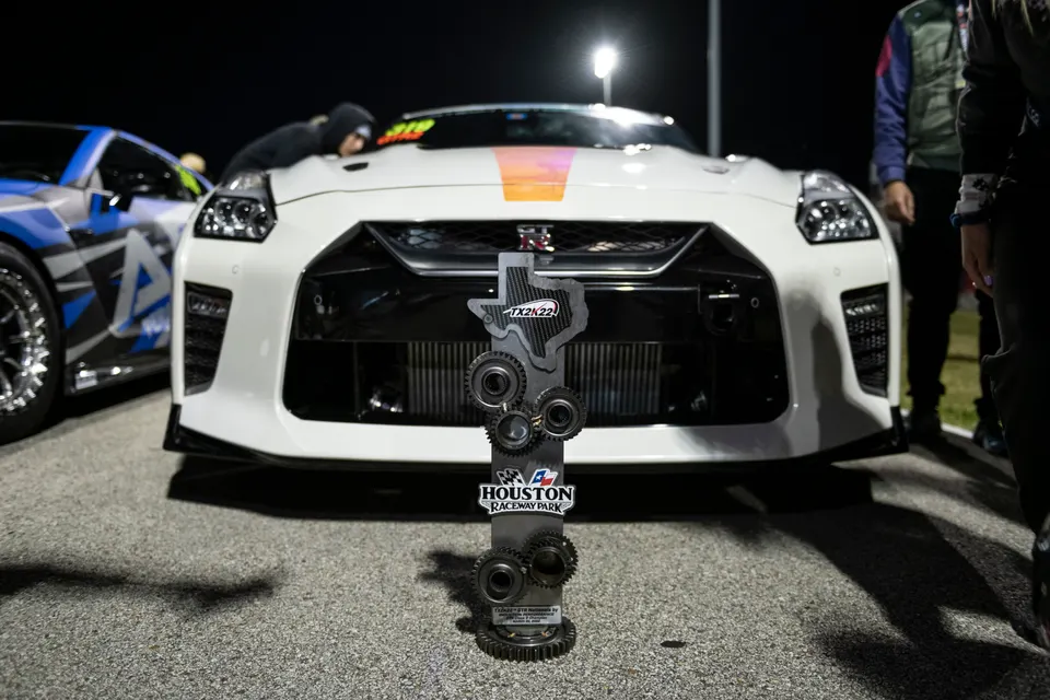 White Nissan GT-R with an orange stripe on the hood, parked at a drag event with a TX2K22 trophy in front.