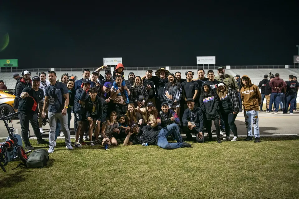 A large group of people, including adults and children, pose for a photo on the grass at a race track at night.