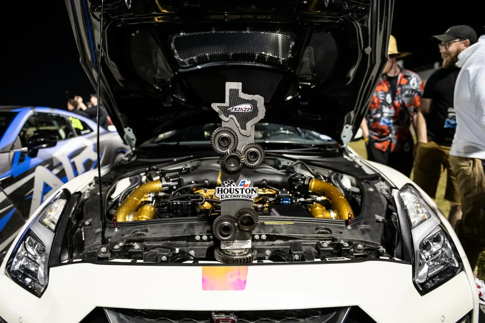 A Nissan GT-R with its hood open, showcasing a custom engine bay with gold piping and a TX2K22 trophy at an event.