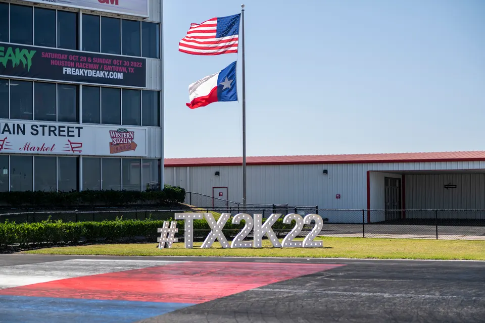 Flags flying over the Houston Raceway Park building with a large #TX2K22 sign on the grass, indicating a track event.