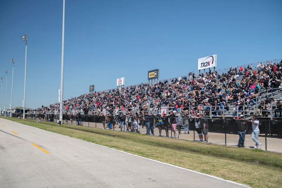 Spectators fill the grandstands at a TX2K motorsports event, with a clear blue sky overhead.
