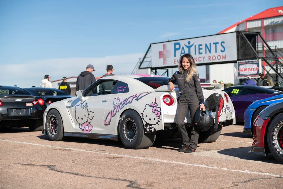 Female driver in racing suit stands next to a white Nissan GT-R with Hello Kitty livery at a drag strip.