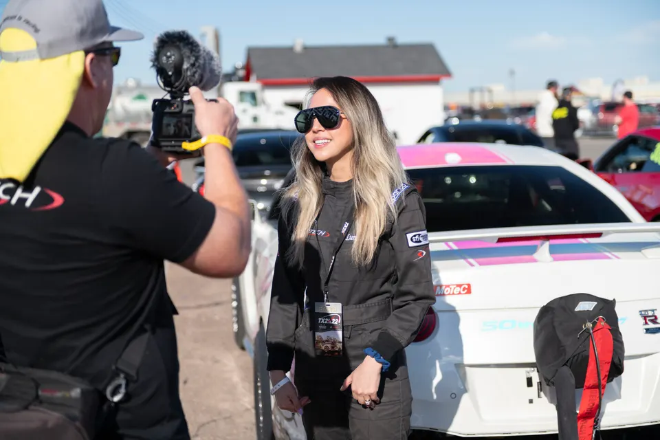 A woman in a racing suit smiles while being filmed by a cameraman at a motorsports event, with a white Nissan GT-R in the background.