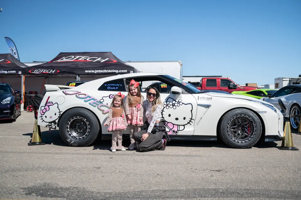 A white Nissan GT-R with Hello Kitty decals, large drag tires, and Jotech Racing branding, with a woman and two children posing next to it at a track event.