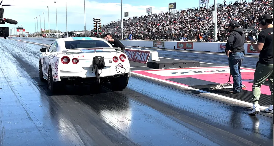 A white Nissan GT-R with a parachute attached at the rear, on a drag strip with spectators in the stands.