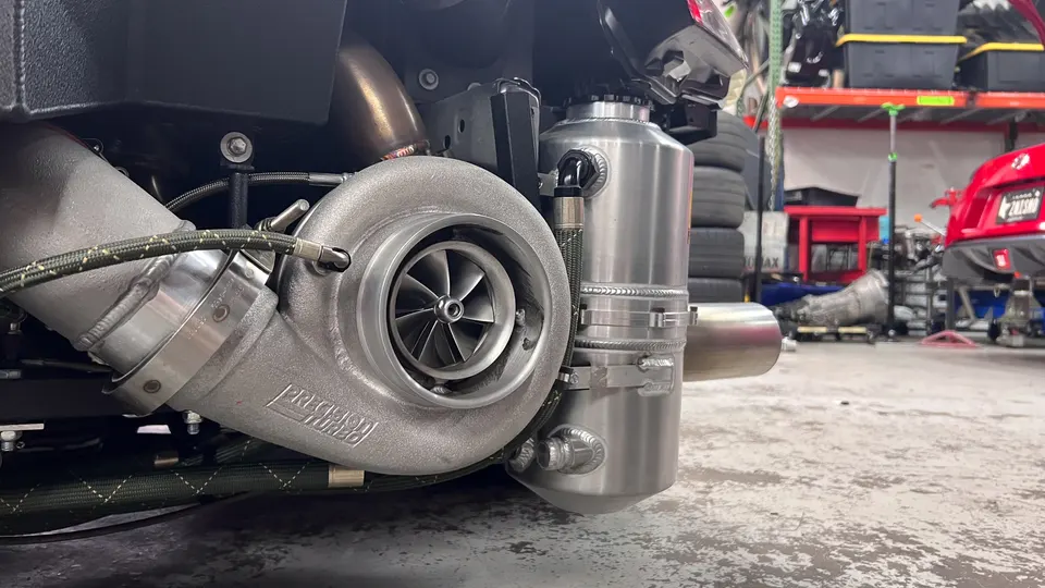 A close-up of a Precision Turbo turbocharger and an aluminum catch can installed in a performance car's engine bay at a tuning shop.