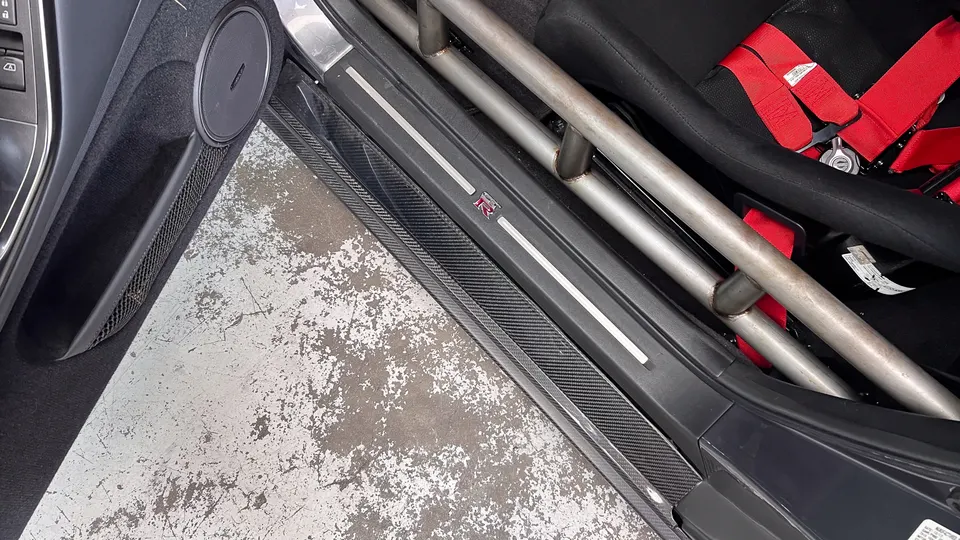 Close-up of a Nissan GT-R interior showing a roll cage, racing seat with red harnesses, and carbon fiber door sill.