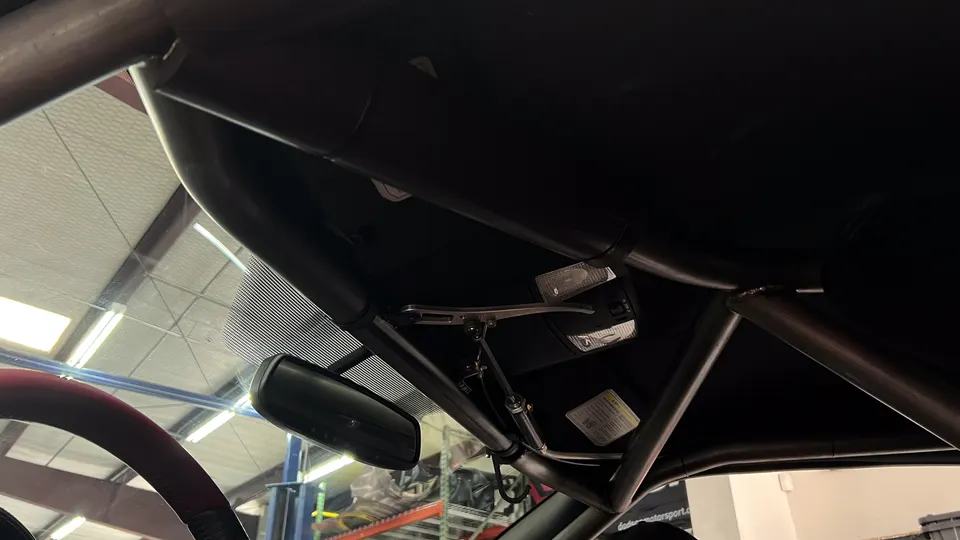 Interior view of a modified car showing a custom roll cage, rearview mirror, and windshield in a shop setting.