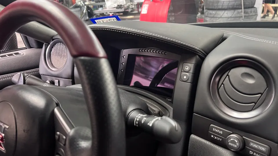 Close-up of a Nissan GT-R interior showing the steering wheel, dashboard, and an aftermarket digital display.