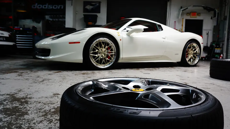 White Ferrari 458 with aftermarket gold wheels in a performance tuning shop, with a loose wheel and tire in the foreground.