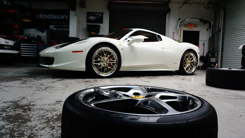 A white Ferrari 458 with custom gold wheels parked in a performance tuning shop with a loose wheel in the foreground.