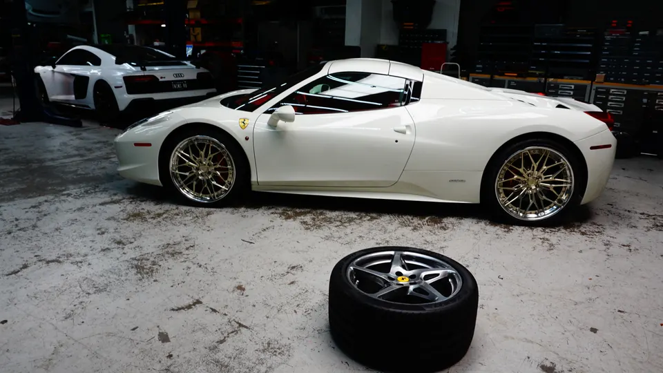 White Ferrari 458 and Audi R8 in a performance tuning shop with aftermarket wheels on the Ferrari and a loose wheel on the floor.