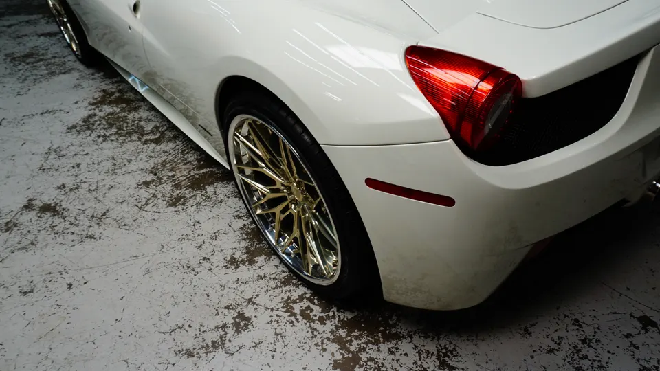A white Ferrari with custom gold wheels and a red taillight, parked on a concrete floor.