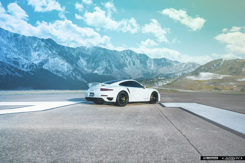 White Porsche 911 Turbo S with black wheels and yellow calipers parked on an asphalt road with snow-capped mountains in the background.