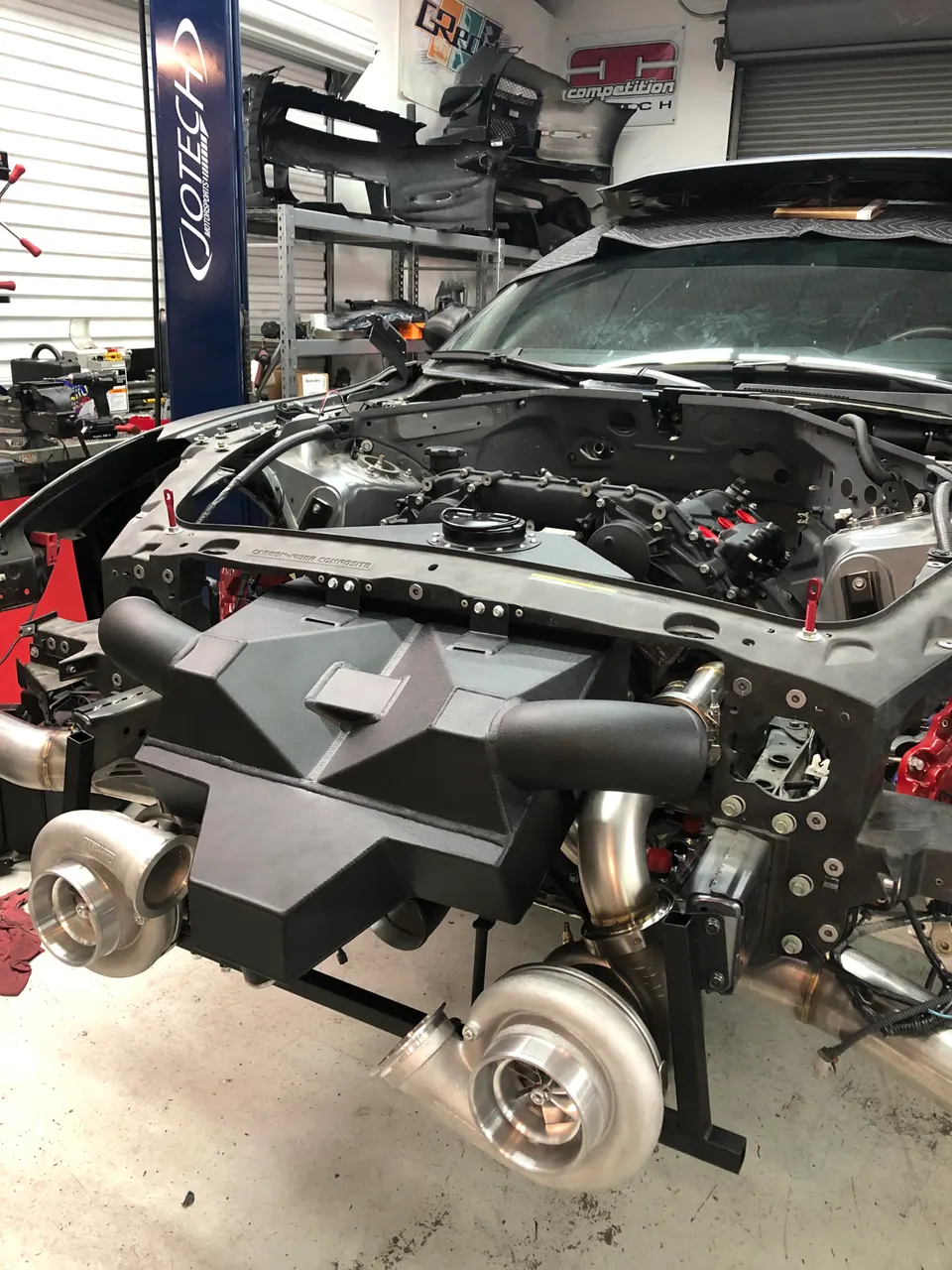 Front view of a high-performance car in a Jotech Motorsports shop, showcasing a custom twin-turbo setup and intake manifold.