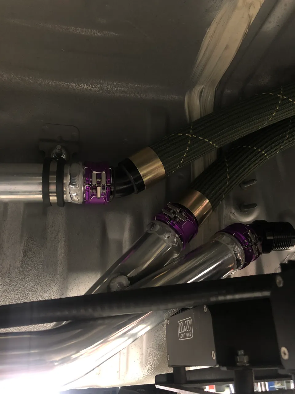 Close-up of custom braided lines, polished pipes, and an M&W Ignitions module in a performance car.