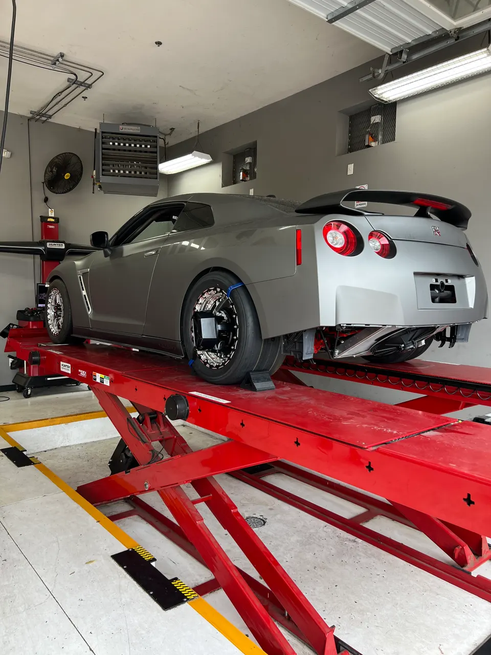 A gray Nissan GT-R R35 on a red alignment lift in a performance tuning shop, with specialized wheels and sensors.