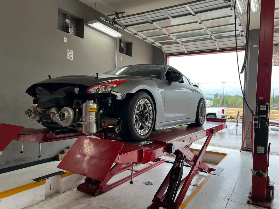 A silver Nissan GT-R on a red lift in a shop, with its front bumper removed revealing a twin-turbo setup.