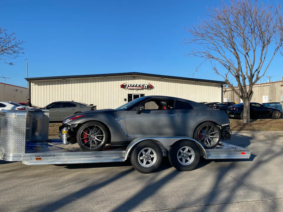 A gray Nissan GT-R on a car hauler trailer parked in front of the Jotech Motorsports shop building.
