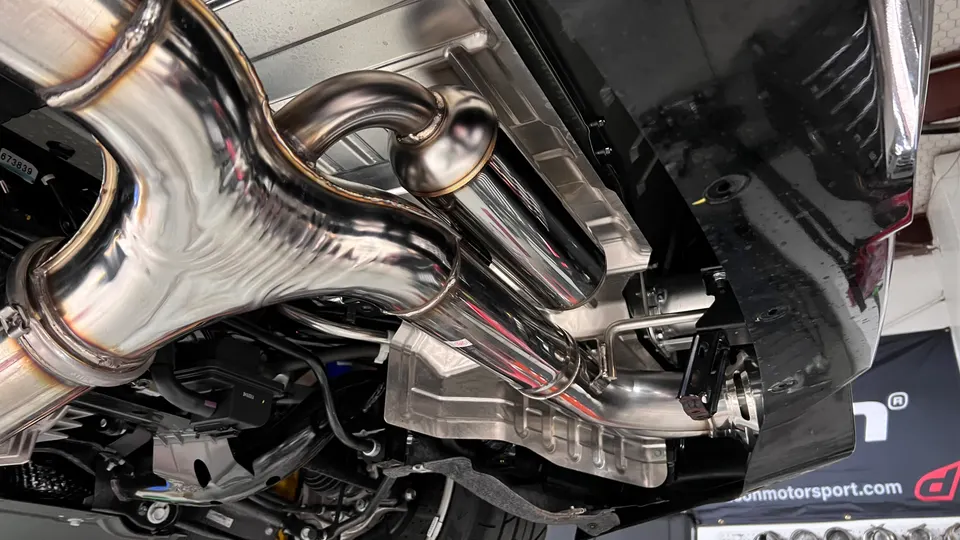 Close-up of a shiny stainless steel performance exhaust system installed on the underside of a car in a shop.